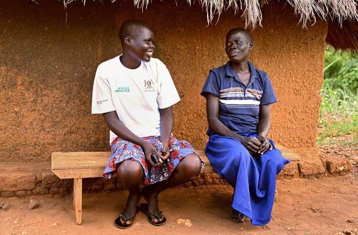 Annet Luka (at left), a South Sudanese refugee, shares a light moment with her mother outside their home in Terego, northern Uganda. Annet has already helped her family by using her new woodworking skills to make the wooden bench that they are sitting on.